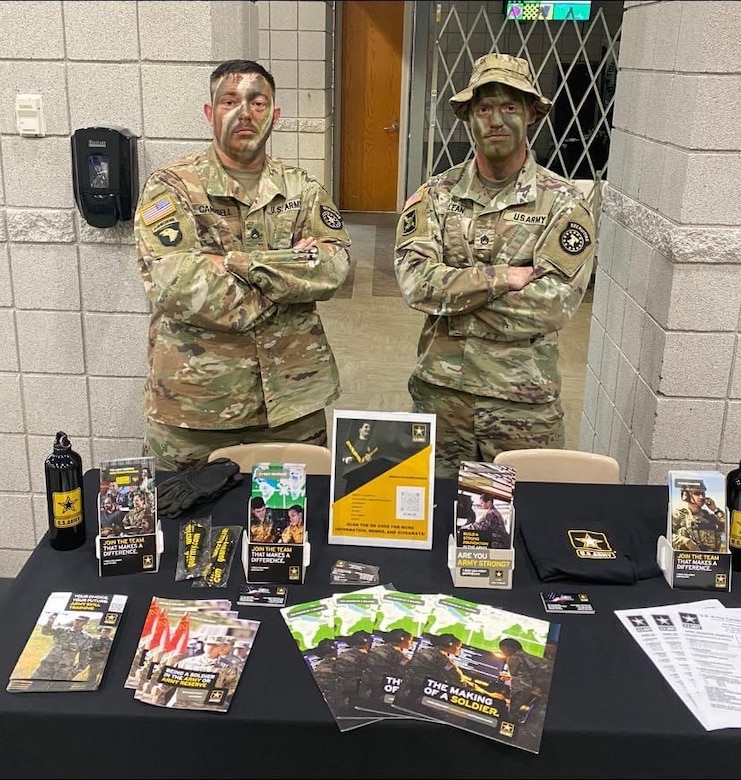 Two men in Army uniforms with camouflage on their face pose behind a table filled with Army branded items