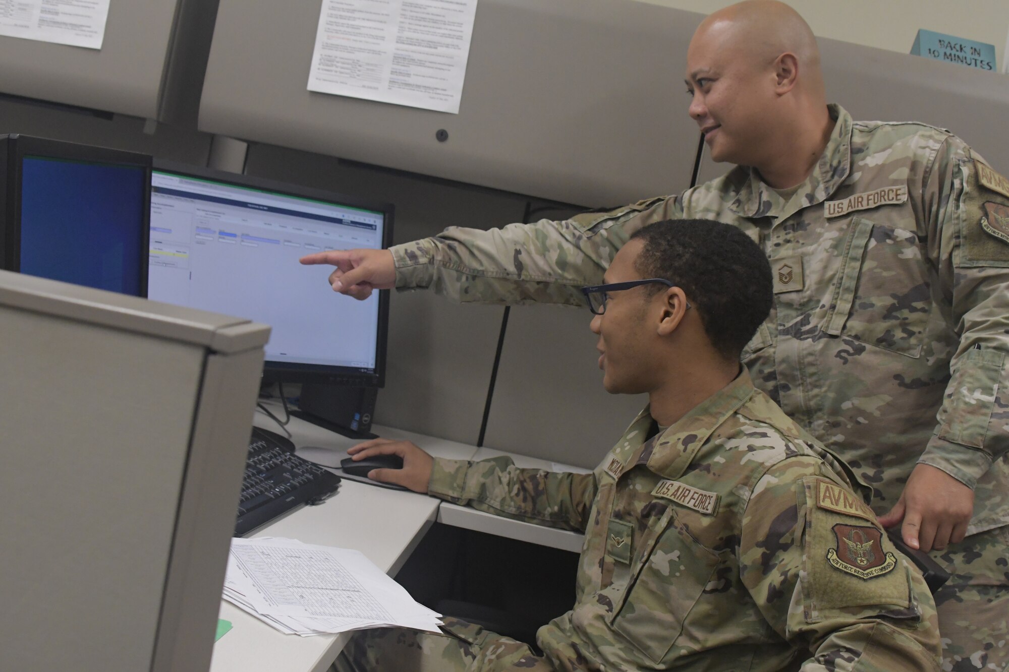 a man wearing a military uniform sits at a computer while another man wearing a military uniform points over his shoulder