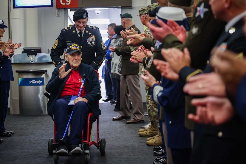 A service member pushes a veteran in a wheelchair through an airport as other service members applaud.