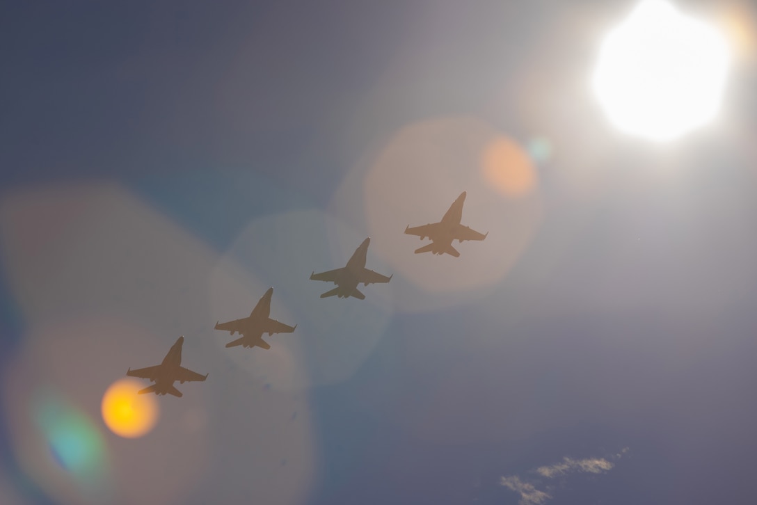 Four military aircraft fly against a blue sky with a bright sun.