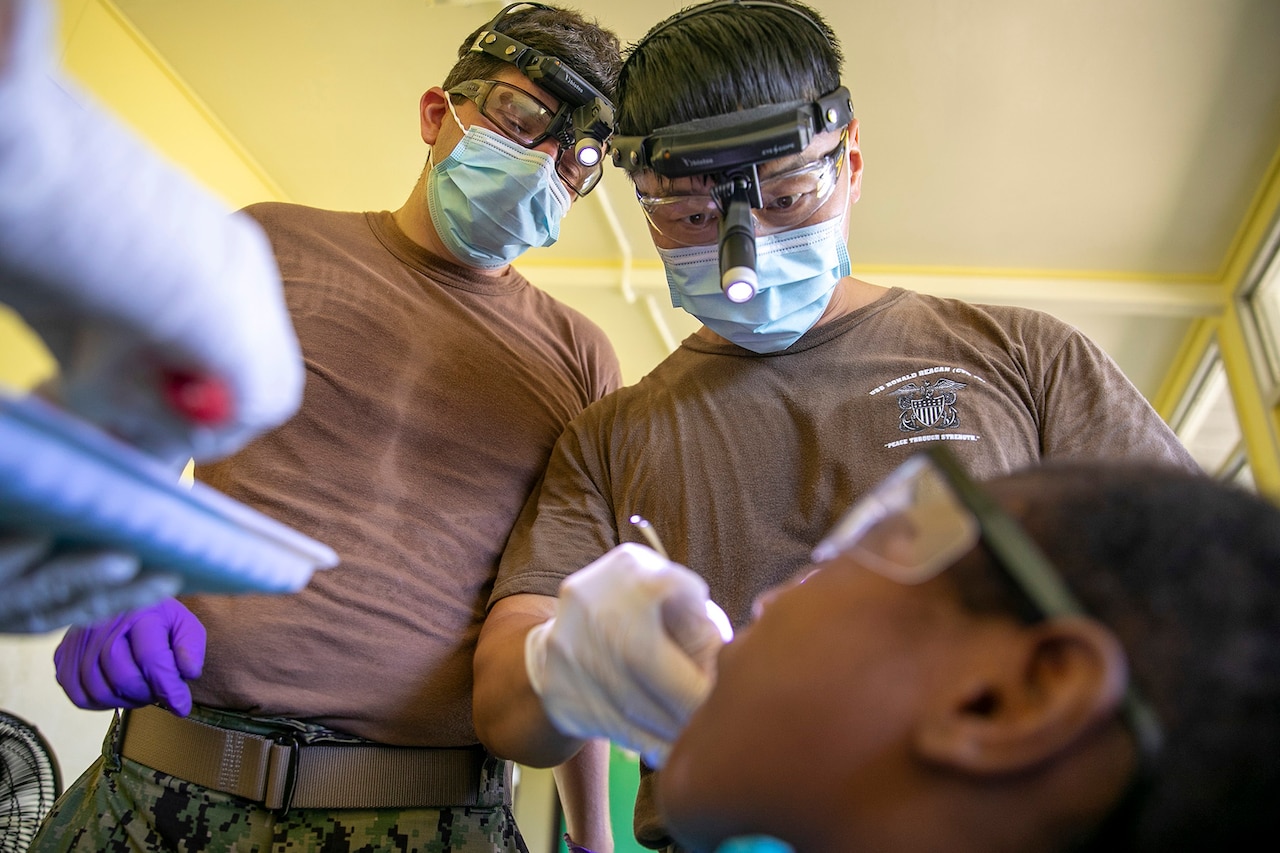 Sailors wearing surgical masks and dental headlights perform a tooth extraction on a child.