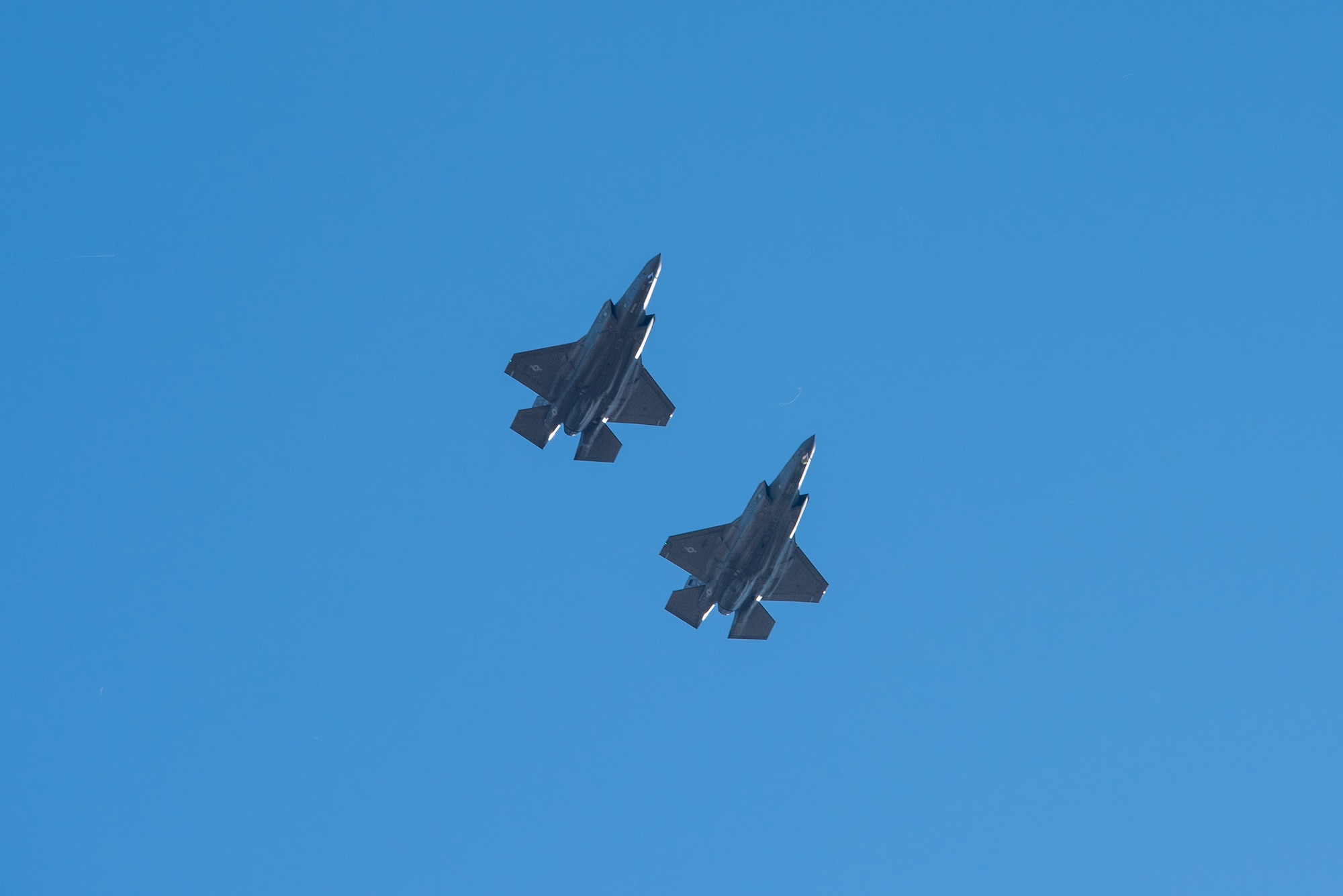 Image of two F-35A fighter jets in formation. looking up at the bottom of the aircraft with blue sky as background.