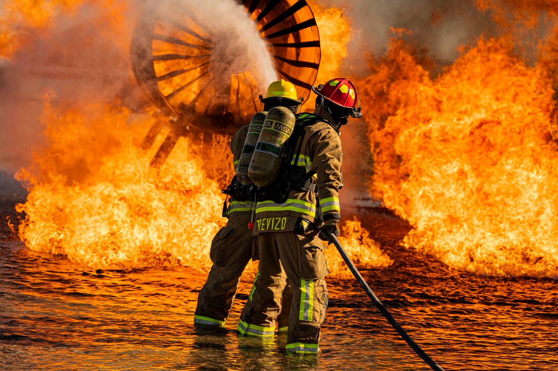 Two airmen in fire protective gear use a water hose on an aircraft fire.