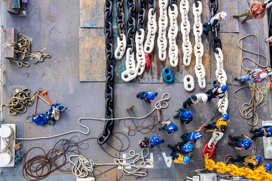 An overhead look at sailors standing next to large chains on the deck of a ship.