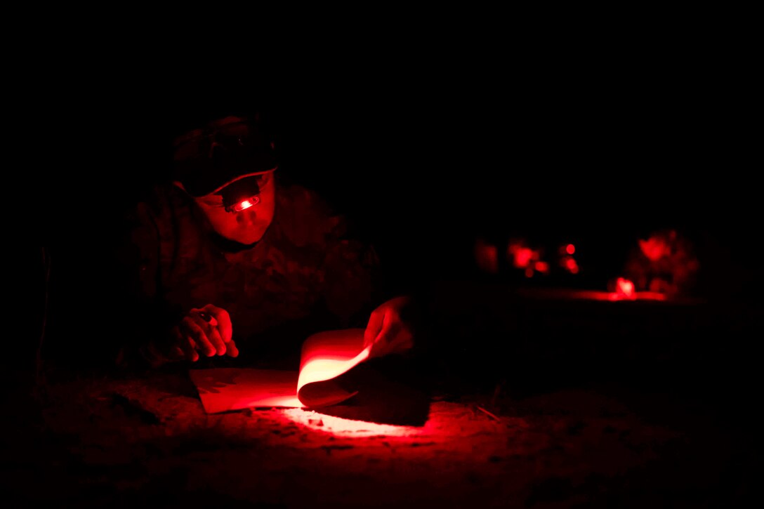An airman illuminated by red light uses a headlight while flipping through papers.