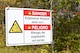 A danger sign is seen hanging on a barbed wire fence in the foreground. The sign has an icon and English and Spanish text warning of explosive hazards. The blurry background contains trees.
