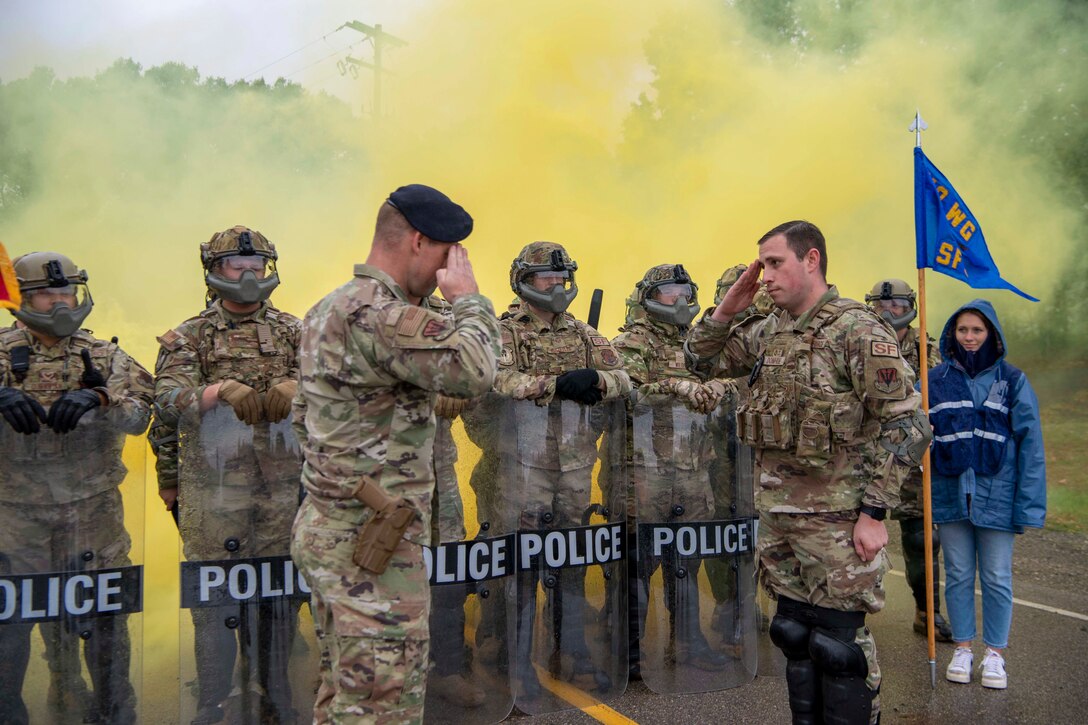 Two service members salute while facing each other as fellow service members wearing tactical gear stand in formation holding shields surrounded by yellow smoke.