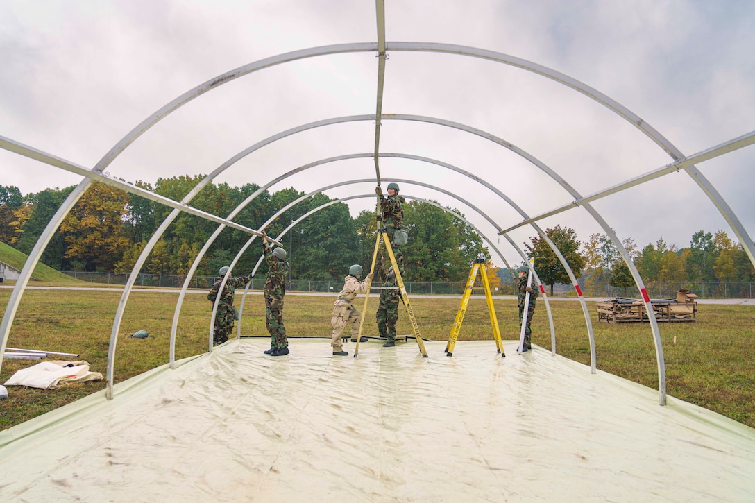 National Guardsmen stand on ladders to put metal materials in place while building a structure in a field with trees in the background.