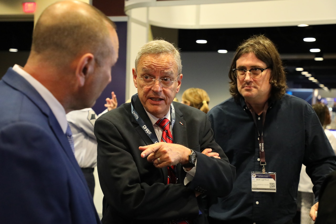 A man in front in business attire talks with two men in a room filled with people.