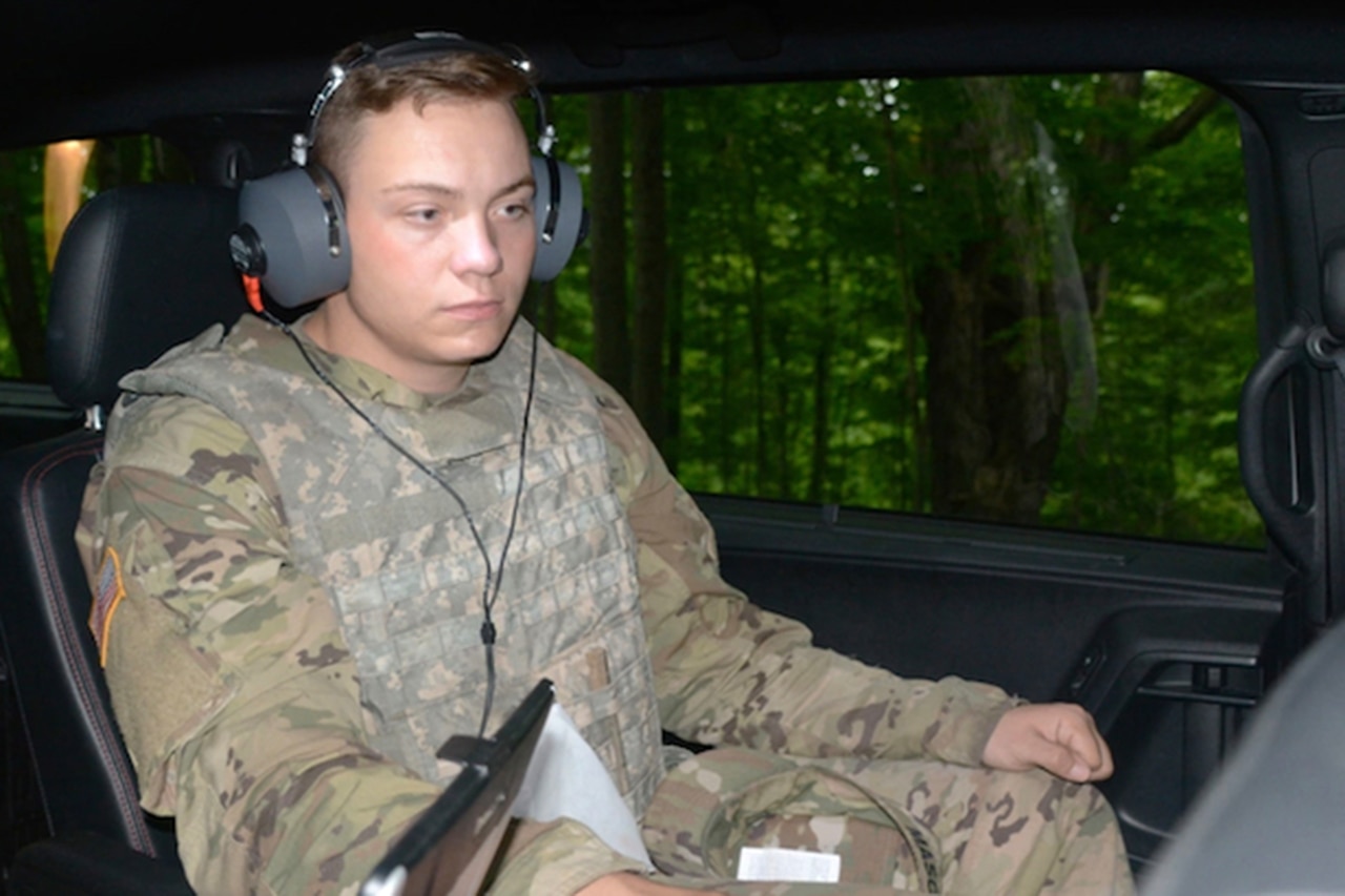 A uniformed service member sits in a vehicle while wearing over-the-ear hearing protection.