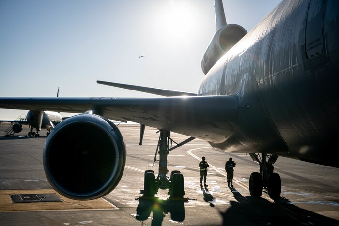 the sun is bright in the center top of the image while there is a large grey military aircraft taking up most of the photo while there are two people in the center lower part of the photo