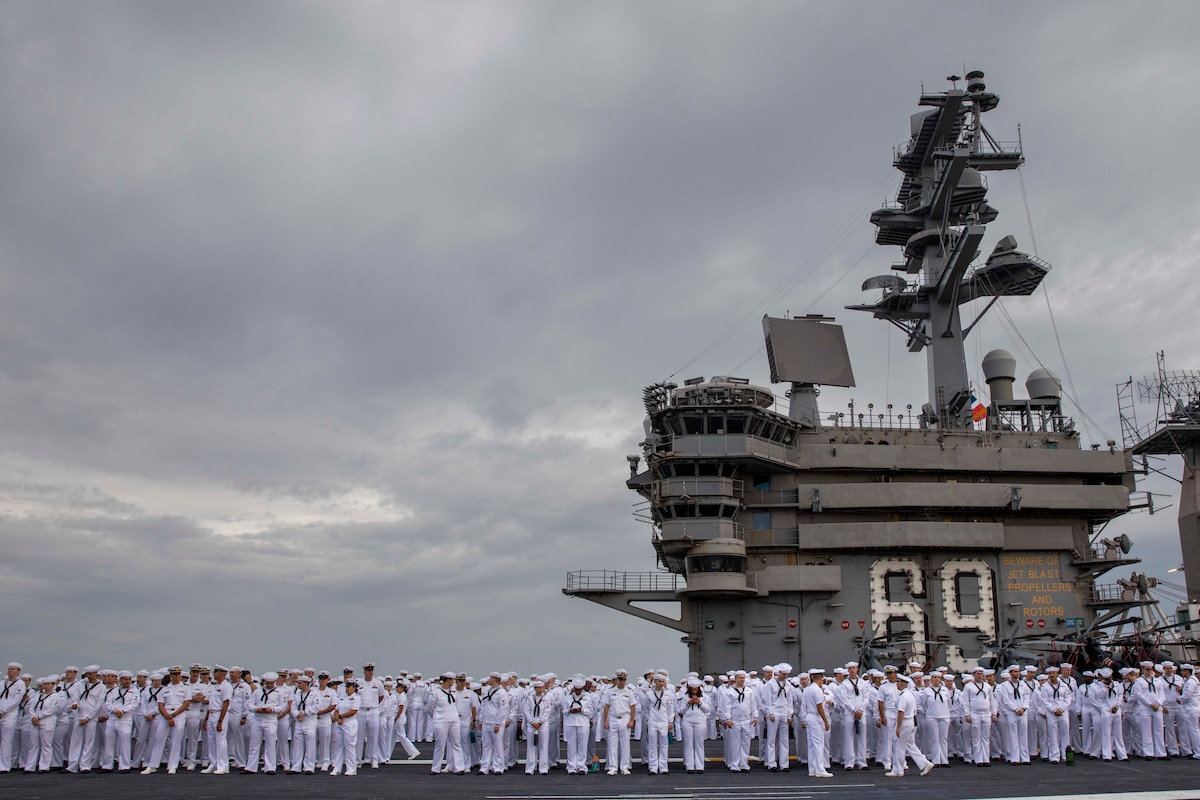 Sailors in uniform stand on the flight deck of an aircraft carrier.
