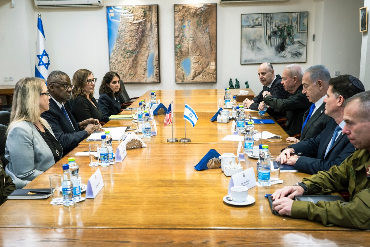 Men and women are seated along each side of a small table with U.S. and Israeli flags.