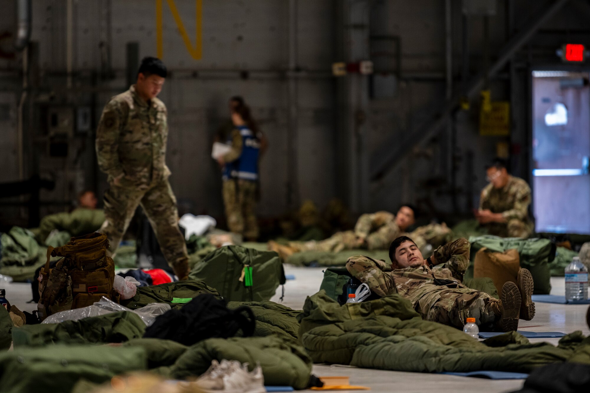 while many sleeping areas of sleeping bags and pads are on the ground in a massive room, a man lays on one and rests