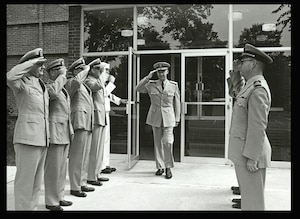 A line of military officers saluting another officer walking out of the door of a building.