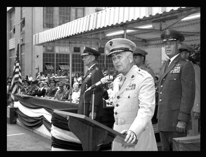 Banner-draped grandstand with seated military and civilian personnel with 4 military officers standing nearby with a podium and micrphone.