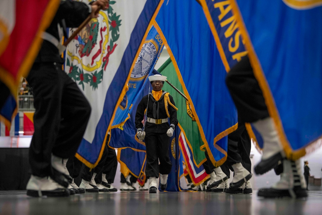 A Navy recruit in a formal uniform marches toward the camera with flags on either side.