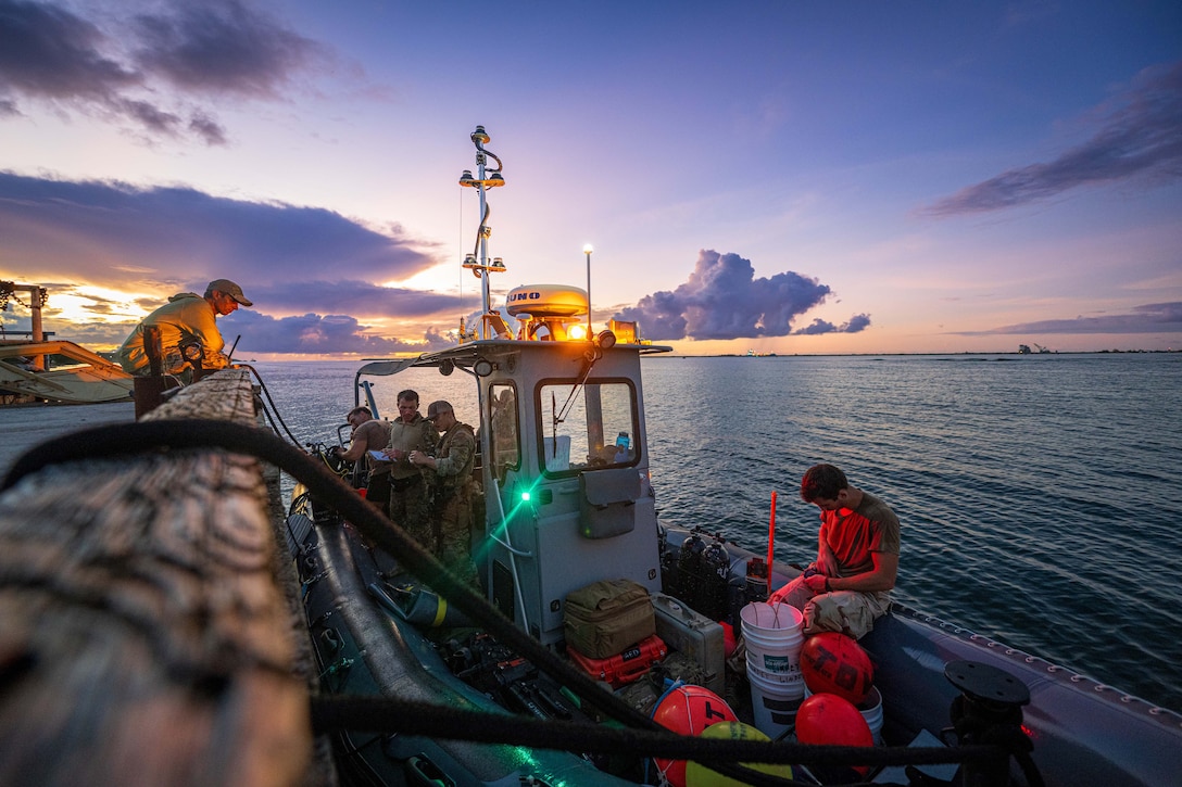 Sailors stand as one is seated on a small boat at sea. The sun is setting in the background.