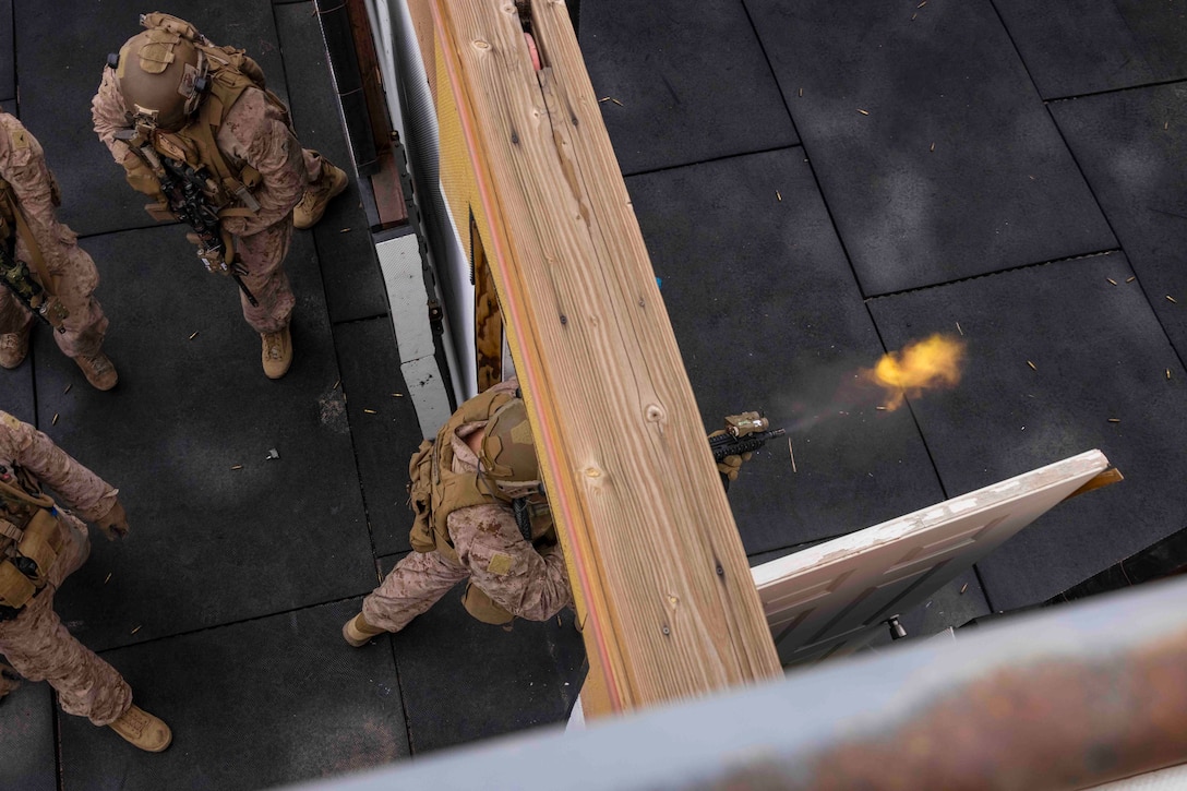 Marines move through a building while one fires a weapon through a door frame.