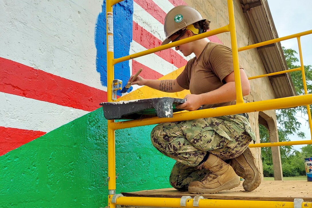 A sailor kneeling on an elevated surface paints a mural on a building.