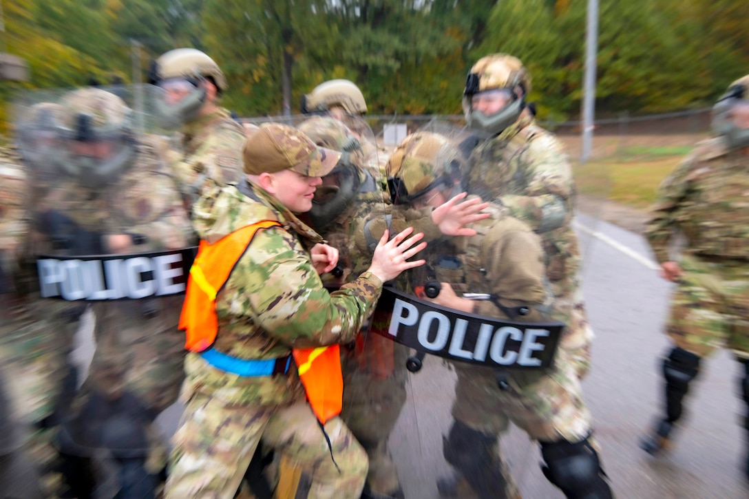 A blurry photo of a service member pushing against fellow service members holding police shields with greenery in the background.