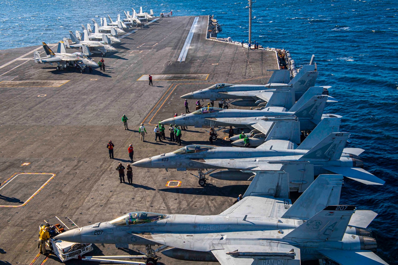 Two rows of aircraft are at the ready on the deck of a military vessel.