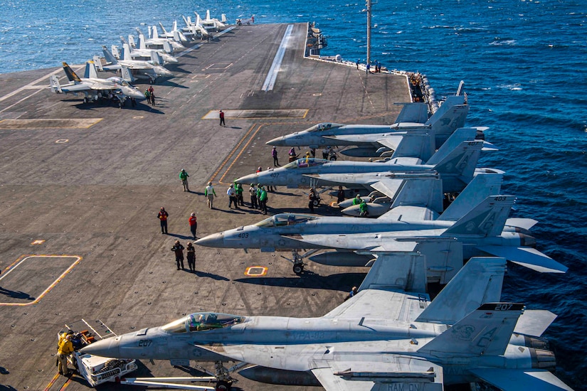 Two rows of aircraft are at the ready on the deck of a military vessel.