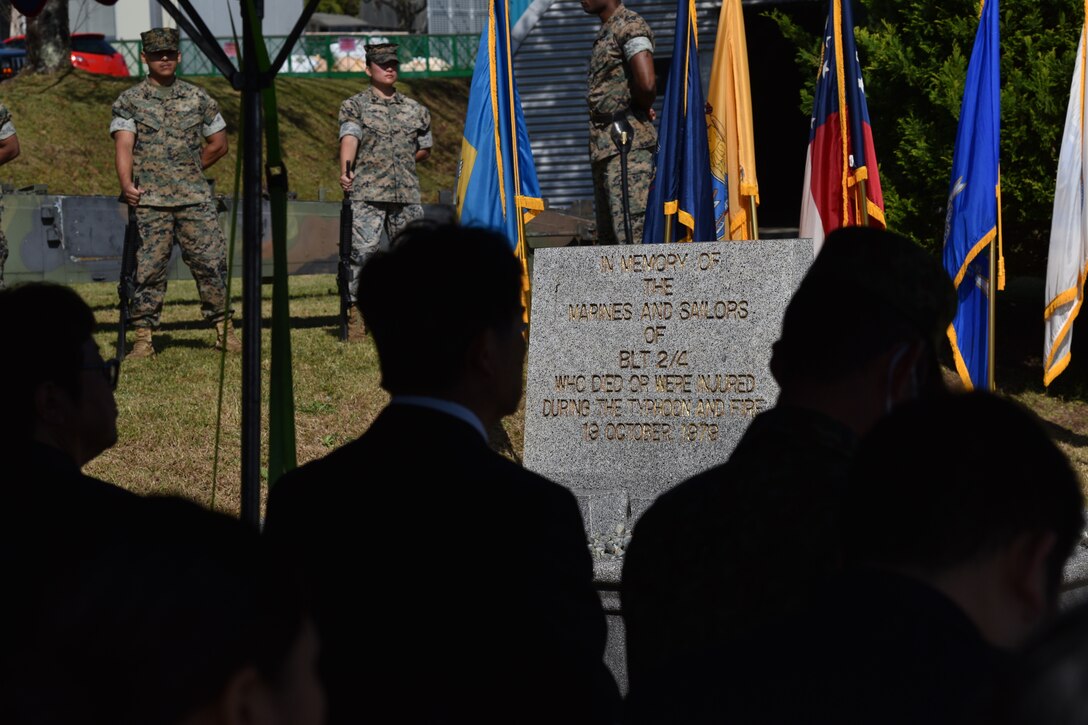 Marines stand in the background, behind the Camp Fuji Fire Memorial Monument for the fallen and injured in the 1979 fire, that occurred during Typhoon Tip. In the foreground, attendees sit in the shade, under a tent. (U.S. Marine Corps photo by Song Jordan)