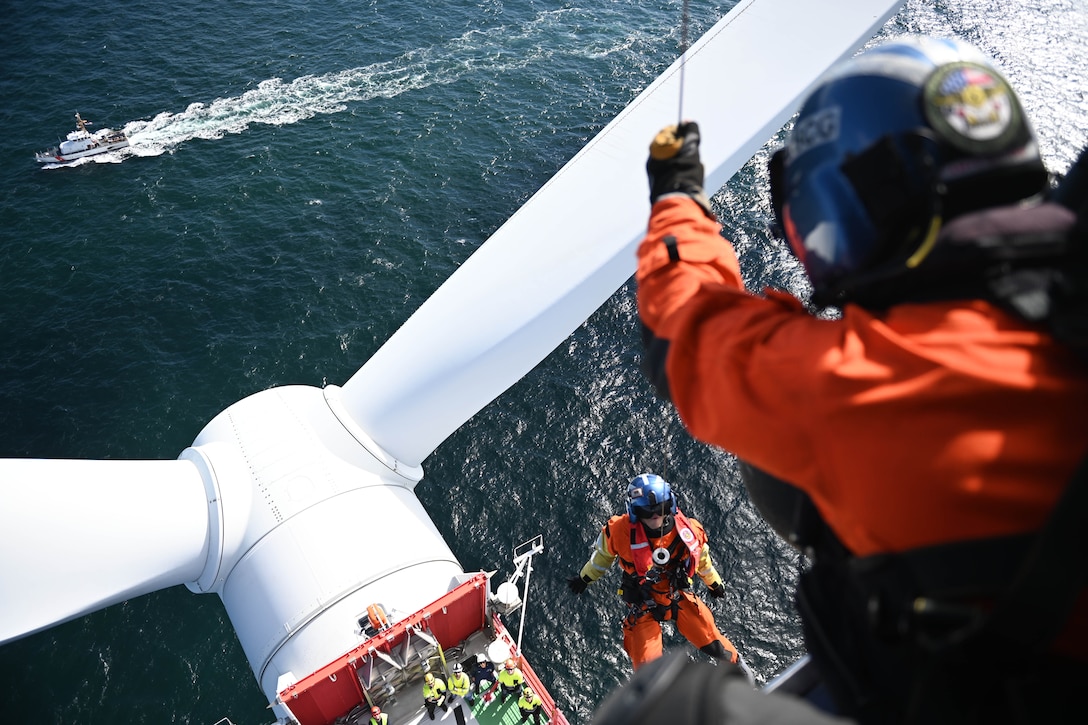A Coast Guardsman is lowered by a cable onto a wind turbine.