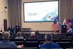 Two men stand in an auditorium presenting information to a large group.