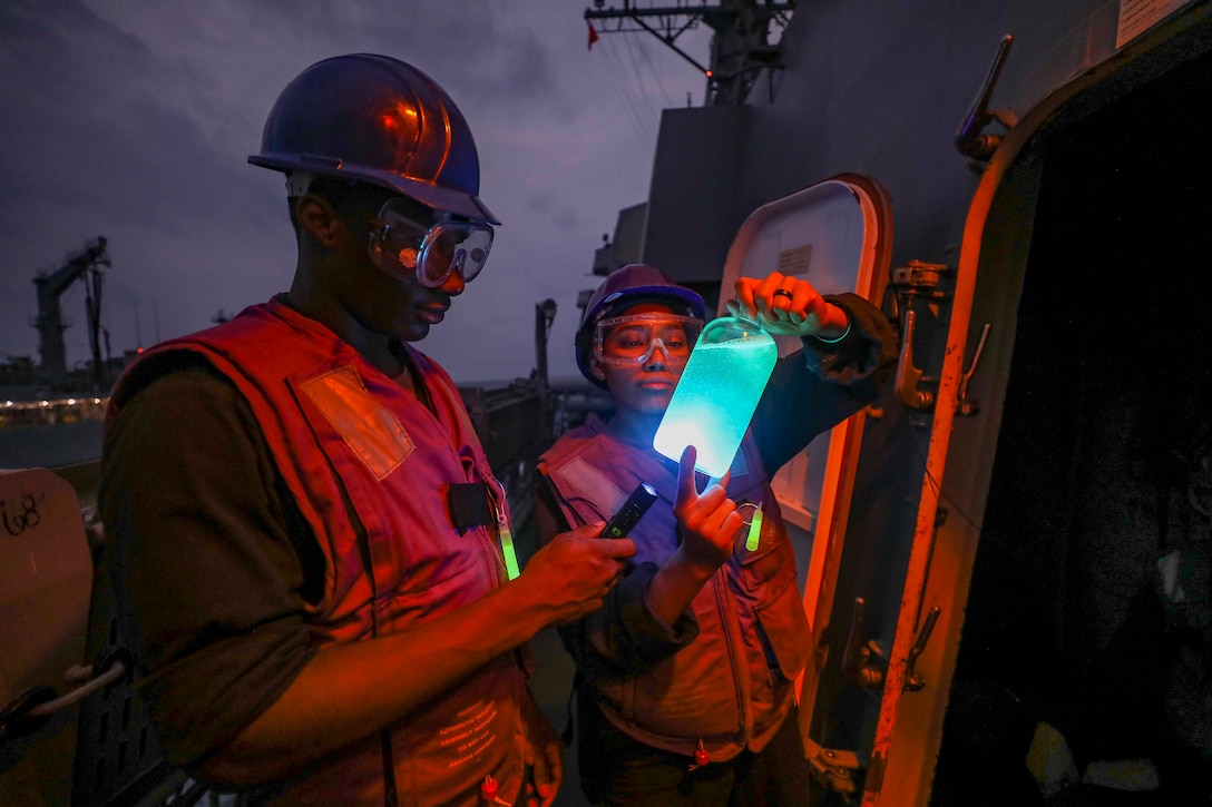 A sailor holds up a fuel sample while another sailor shines a flashlight on the sample. A red light illuminates the ship.