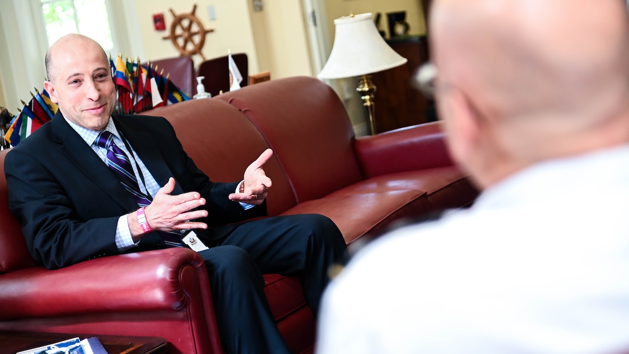 A man in a suit gestures with his hands while seated on a couch.