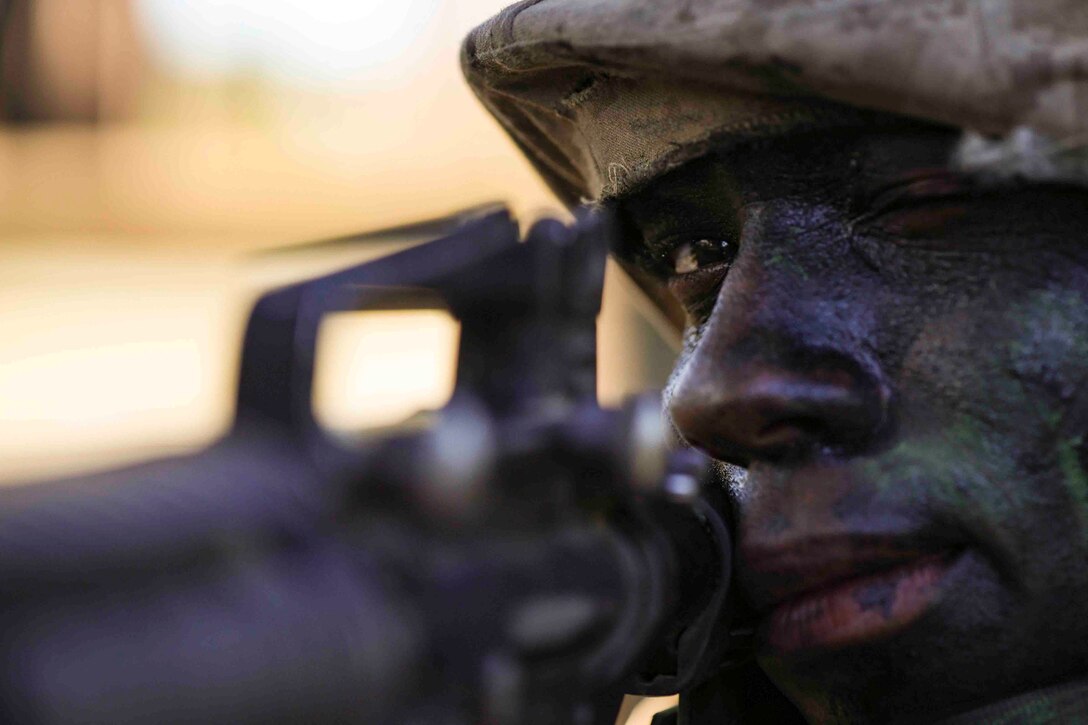 A recruit with camouflage paint on their face looks through the scope of a weapon.