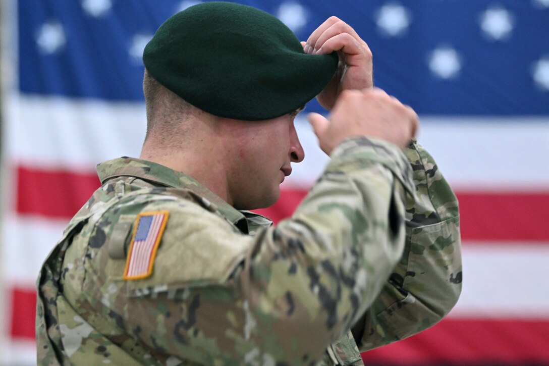 A soldier puts on a green beret with blurred American flag in the background.