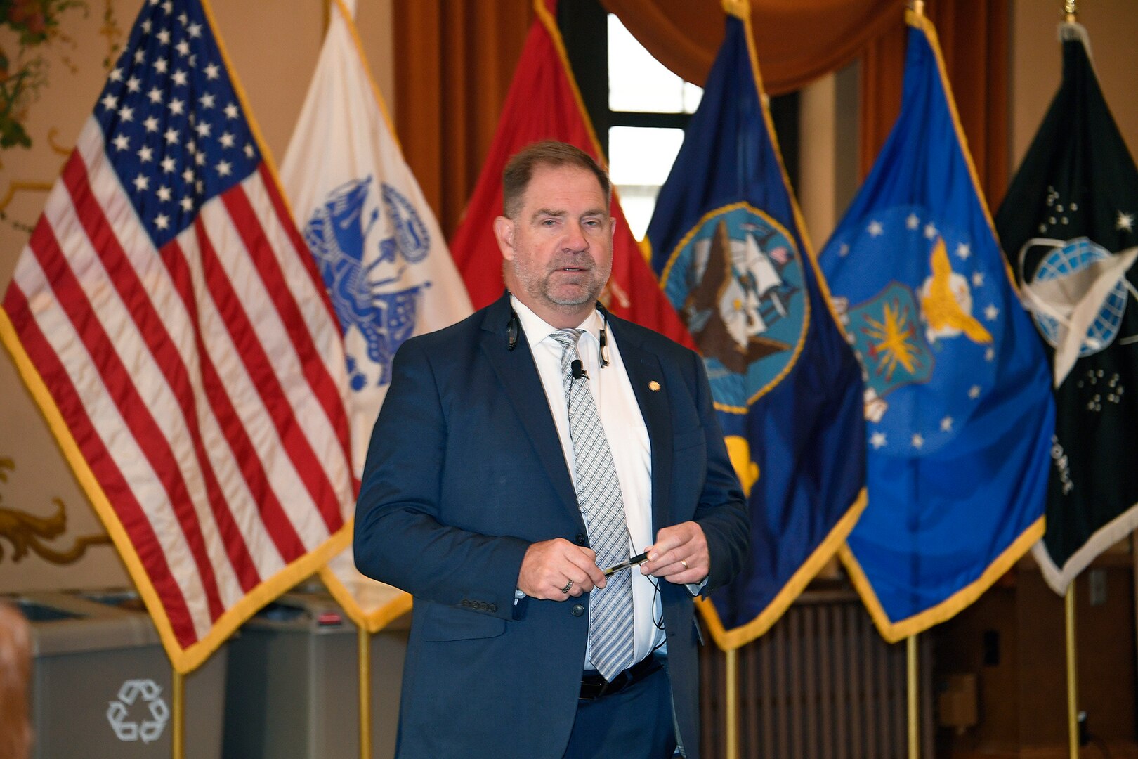 Man in blue suit speaks while holding a pen and standing in front of flags