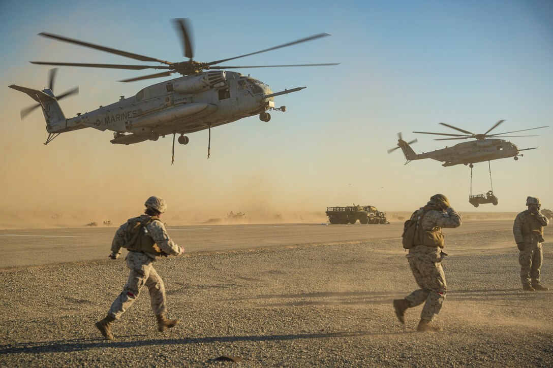 A helicopter carrying a vehicle flies next to another as Marines run in the desert below with more vehicles parked in the background.