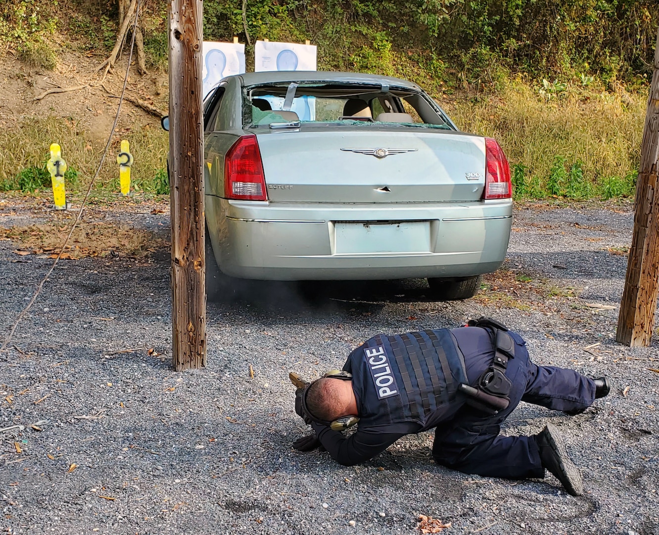 Photo is of a DLA police officer kneeling down on ground pointing weapon at a broken down vehicle during a training event.
