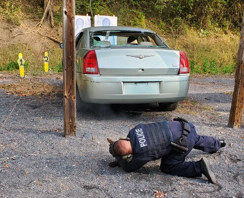 Photo is of a DLA police officer kneeling down on ground pointing weapon at a broken down vehicle during a training event.