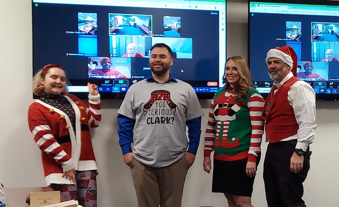 A blonde woman bends over backwards with a ugly Christmas sweater on. Next to heris a man with a Seriously Clark? T-Shirt which is gray and he has it over a blue dress shirt. Next to him is another blonde woman with a Ugly Santa sweater on and a man with a graying beard that has a Santa hat on, a red vest over a white dress shirt in a conference room with screens showing people tuning in virtually.