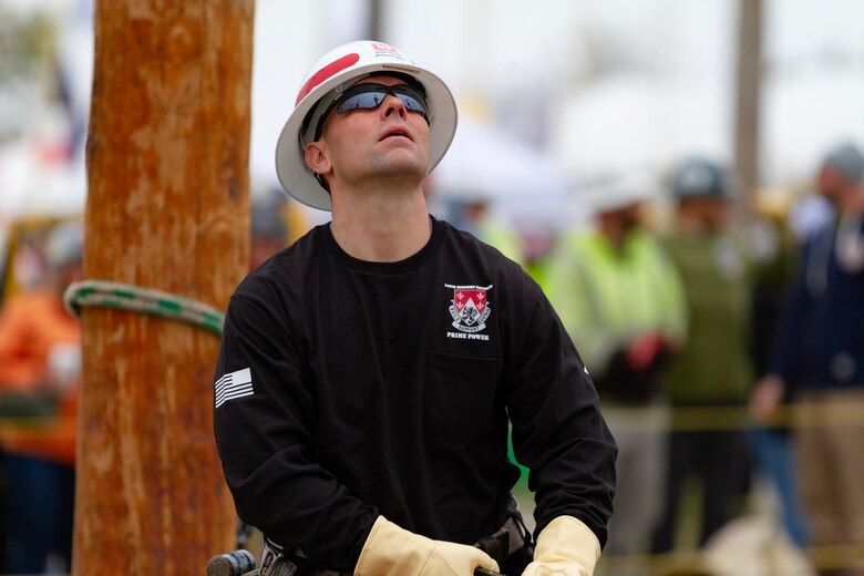 A man in a black shirt with a white hardhat stands in front of a power pole and looks up.