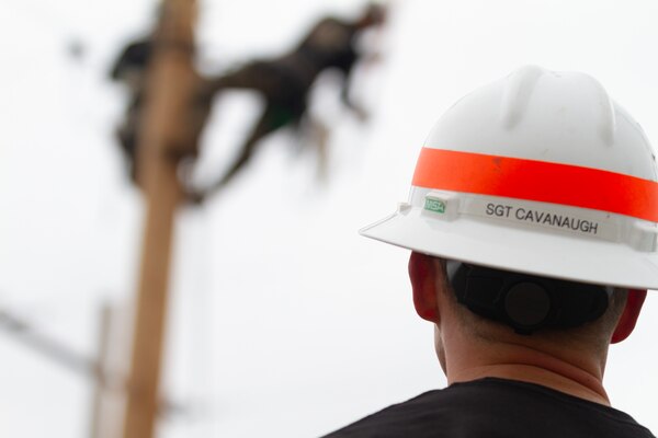 A man in a black shirt and a white hardhat looks up at a utility pole.