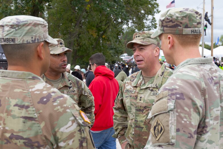 Four men in military uniforms talk with a man in a red sweatshirt in the background.