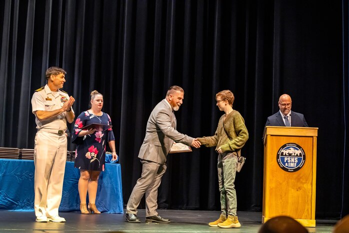 : Capt. JD Crinklaw applauds as Jaime Paylor, machinist, Shop 31, Inside Machinist, receives her certificate from Shop 31 Superintendent Dan Arnall Sept. 29, 2023, during the annual Puget Sound Naval Shipyard & Intermediate Maintenance Facility Apprentice Program graduation ceremony at the Bremerton High School Performing Arts Center, in Bremerton, Washington. (U.S. Navy photo by Jeb Fach)