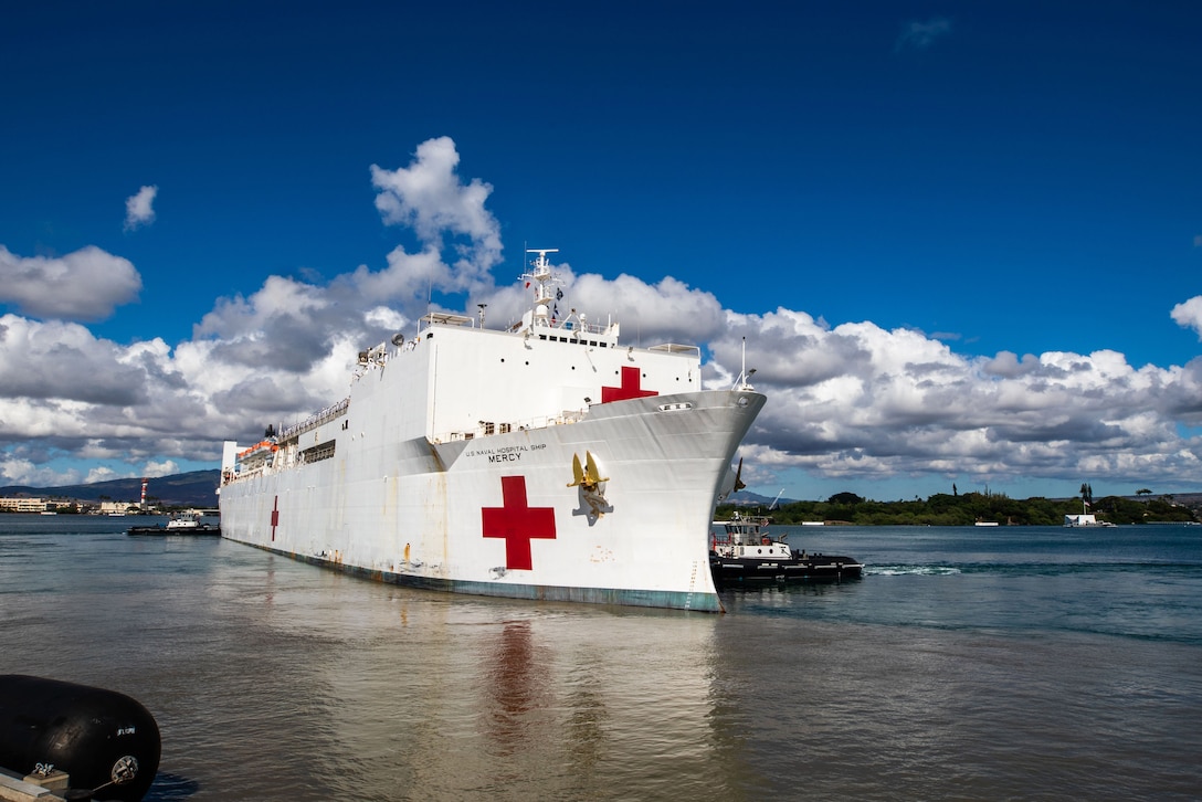 A Navy hospital ship sets sail on a bright day with low-hanging clouds.