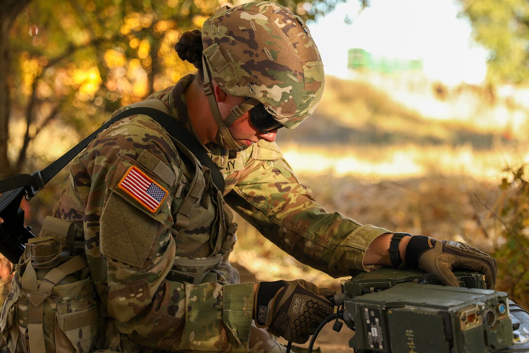 A soldier looks down to check equipment in a wooded area.
