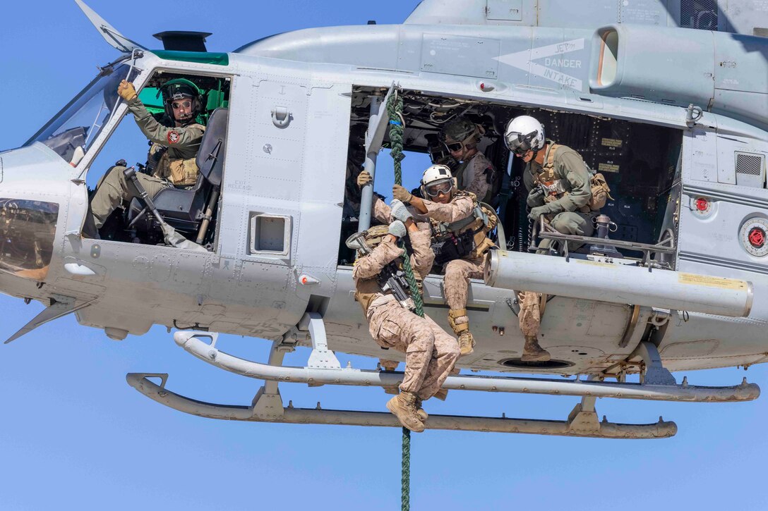 A Marine prepares to fast-rope out of an airborne aircraft as others sit inside the aircraft.