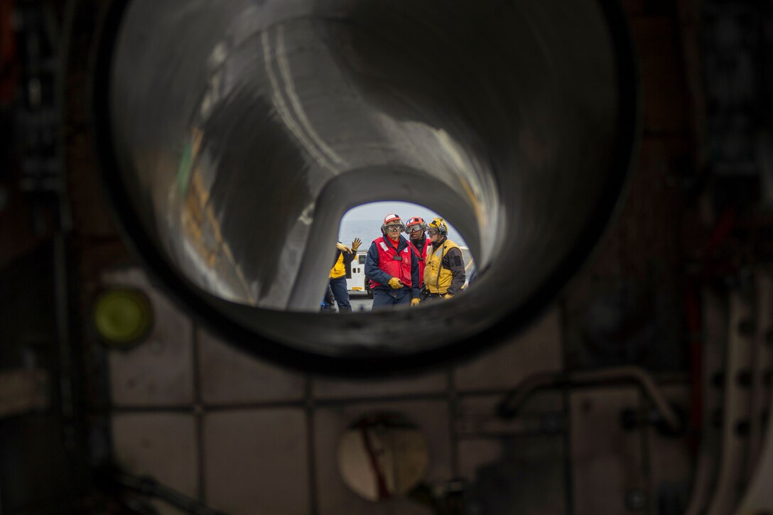 Three sailors stand next to each other aboard a ship at sea as seen through a pipe.