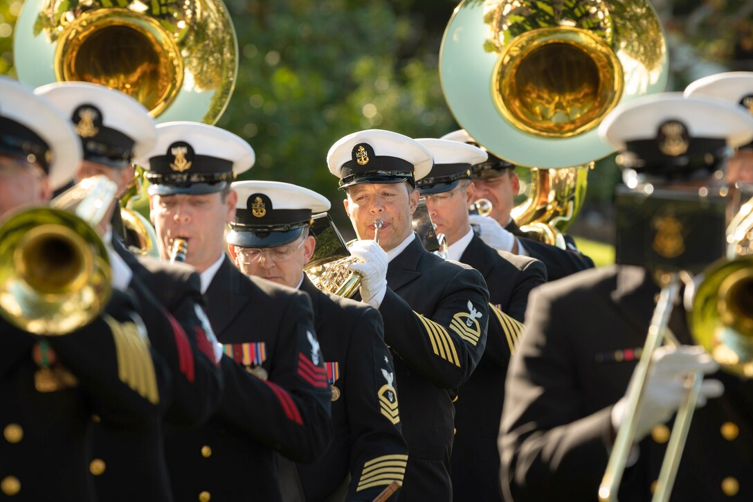Sailors stand in formation while playing instruments with blurred greenery in the background.