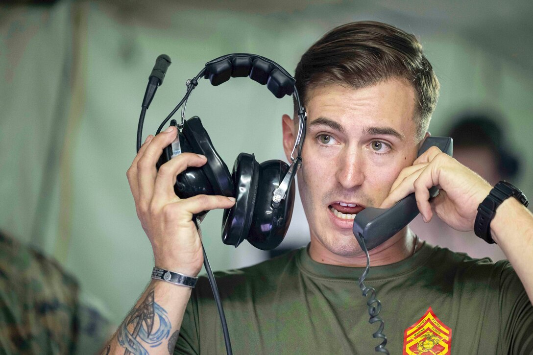 A Marine holds a corded phone to one ear and a headset to the other.