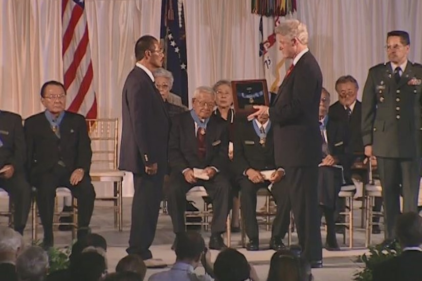 Two people face each other while one holds a plaque. Others sit in rows behind them.